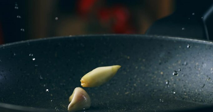 Super Slow Motion Close Up of Garlic Cloves Dropping into Hot Oil Creating a Splash in a Frying Pan with Dramatic Lighting Against a Dark Background at 1000 fps.