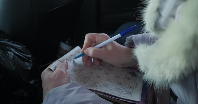A woman in winter clothes, sitting in a moving car, solves a crossword puzzle by writing down the answer. Hand held. Siberia.