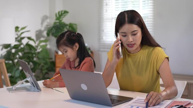 A mother juggles a phone call while helping her daughter with homework, demonstrating multitasking and care in a home setting.