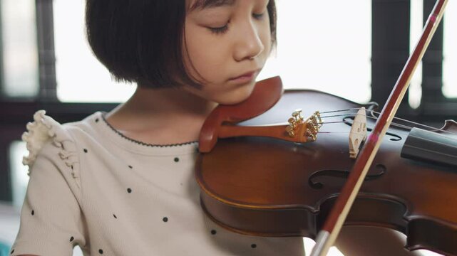 Young Asian girl practices violin in the room, showing talent and passion for classical music. Close-up highlights her focused expression and precise movements with the classical string instrument. 