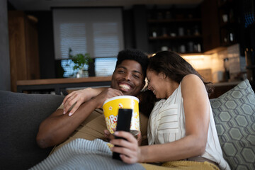 A young couple watching a horror movie at home. The woman looks frightened or grossed out, while the man smiles, enjoying the moment. They are sitting on a cozy couch with popcorn