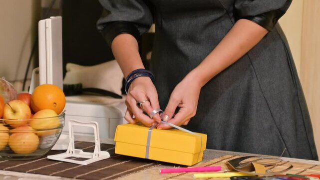 A woman wraps a gift in a yellow gift box. The scene focuses on the act of gift-wrapping, showcasing the care and attention to detail involved in preparing a present.