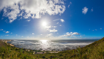 A bright sun shines over a vast ocean with rocky shores and breaking waves under a blue sky with scattered clouds