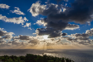 The sun breaks through dramatic clouds over a calm ocean, casting a golden light on the water.