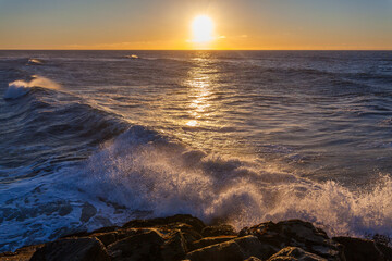 Obraz premium Waves crash against a rocky shoreline at sunset, with the sun setting over the ocean horizon.