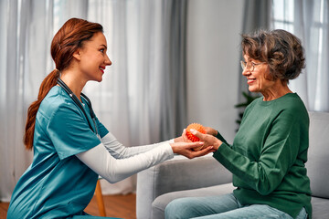 Adult nurse physiotherapist helps elderly woman to do exercise with spiky ball in hands. Restoring...