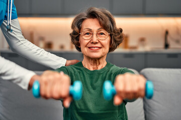 Portrait of a beautiful smiling elderly woman holding light dumbbells and smiling. Nurse helping...