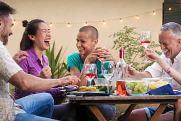 Group of diverse mature friends enjoying food and drinks, laughing together at a backyard dinner party