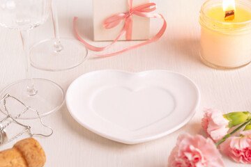 Empty heart shaped festive ceramic plate served on white wooden table with wine glasses, pink carnation flowers, gift box and glowing candle light for romantic engagement proposal or valentines day 