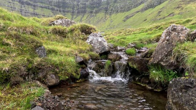 Pure mountain stream flows over rocky terrain through the majestic green mountains. Rain falls over this amazing landscape. Located on the island of Kunoy in the Faroe Islands, Kingdom of Denmark.