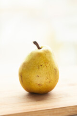 A single ripe pear rests on a light wooden surface, bathed in soft, natural light.  Its yellow skin shows subtle markings.
