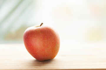 A single, ripe red apple sits on a light wood surface, bathed in soft, natural light.  A simple, healthy image.