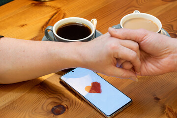 The image shows a couple sitting at a table with two cups of coffee. Their hands are clasped together, and there is a smartphone between them with a red heart on the screen.