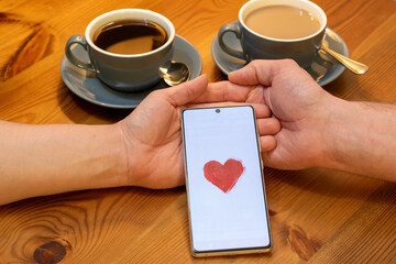 The image shows a couple sitting at a table with two cups of coffee. Their hands are clasped together, and there is a smartphone between them with a red heart on the screen.