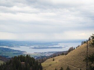 Fototapeta premium A breathtaking view of Chiemsee lake as seen from a hillside in Bavaria, Germany, with surrounding fields, forests, and distant islands under a cloudy sky.
