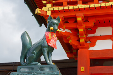 Kyoto, Japan - Sep 23 2024, Close-up view of the stone statue of kitsune, a Japanese fox holding a golden object in its mouth, with red facade of Fushimi-Inari Temple in background, Kyoto, Japan