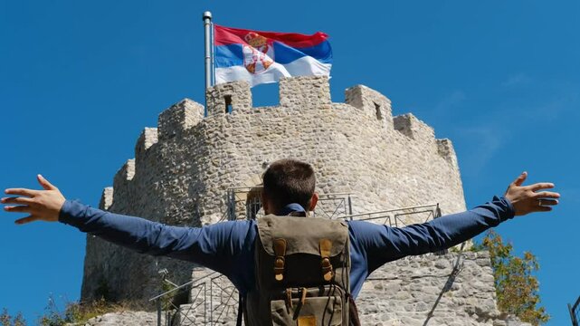 A man stands near the castle with arms raised as if embracing whole world. A popular historical place among tourists. Old Town Fortress in Uzice, Serbia. Castle with Serbian flag on rocky mountain.
