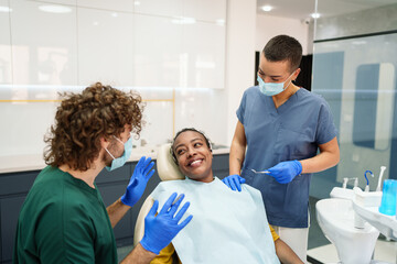 A Black female patient smiles in a modern dental clinic as two healthcare professionals, wearing...