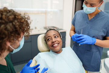 A Black female patient sits comfortably in a dental chair, sharing a warm smile with her diverse dental team. The professionals, wearing protective masks and gloves.