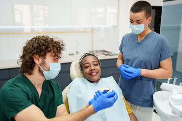 Fototapeta premium A Caucasian male dentist consults with a smiling Black female patient in a modern dental office. A female dental assistant observes the interaction, emphasizing a professional and caring environment.