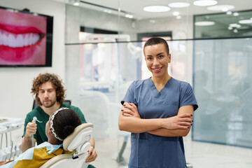 Smiling Caucasian female dental assistant in a contemporary clinic, wearing a blue scrub. A diverse team works on a patient, ensuring comfort. The setting is bright and professional.