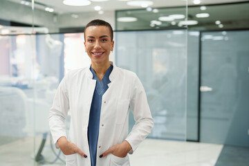 Fototapeta premium A smiling Caucasian female dentist stands confidently in a modern dental clinic, wearing a white coat over blue scrubs. The bright, professional environment underscores her welcoming demeanor.