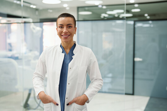 A smiling Caucasian female dentist stands confidently in a modern dental clinic, wearing a white coat over blue scrubs. The bright, professional environment underscores her welcoming demeanor.