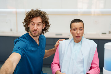 A concerned Caucasian male dentist in blue scrubs attends to an uncomfortable Caucasian female...