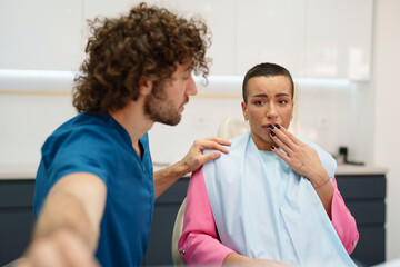 Obraz premium A Caucasian male dentist consoles an uncomfortable Caucasian female patient in a modern dental clinic. The dentist, dressed in blue scrubs, reassures the patient, who is seated in the dental chair.