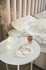 A cozy bedroom scene with a cup of coffee, candle, glass of water, accessories for make up on whet table nearest the bed.