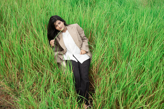 a young Indian female model with black hair, wheatish skin, Indian features wearing a brown coat over a white t shirt and black trousers posing middle of an open grass field