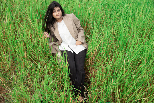 a young Indian female model with black hair, wheatish skin, Indian features wearing a brown coat over a white t shirt and black trousers posing middle of an open grass field