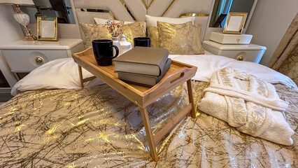 Luxurious Bedroom Setting With Wooden Breakfast Tray, Books, And Coffee Mugs On A Bed With Elegant Gold-Patterned Bedding, Concept Of Comfort And Relaxation, Ideal For Interior Design And Hospitality.