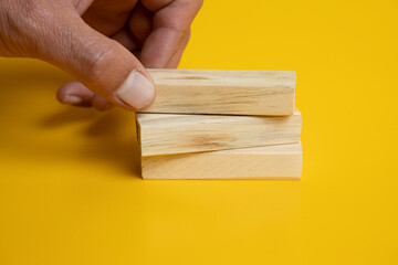 placing a wooden Jenga blocks on two more blocks arranged neatly on a solid yellow background.