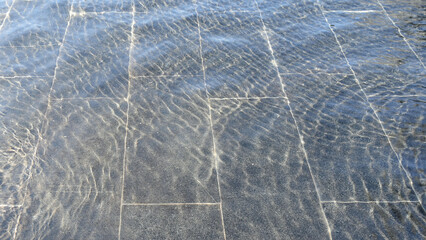 A thin layer of water with ripples on dark marble fountain tiles, illuminated by sunlight. Texture in full screen.