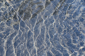A thin layer of water with ripples on dark marble fountain tiles, illuminated by sunlight. Texture in full screen.