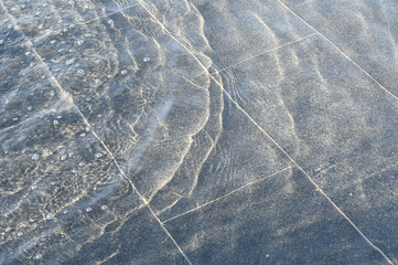 A thin layer of water with ripples on dark marble fountain tiles, illuminated by sunlight. Texture in full screen.