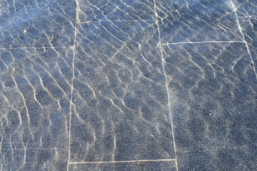 A thin layer of water with ripples on dark marble fountain tiles, illuminated by sunlight. Texture in full screen.