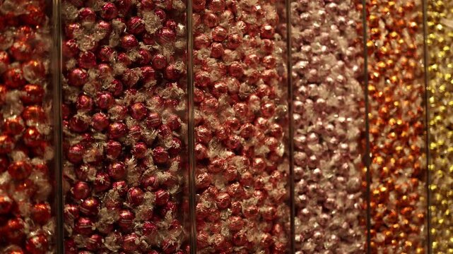 Rows of luxurious wrapped chocolates in shades of red, pink, orange, and gold fill the display in a candy shop. The rich colors and shimmering packaging create an atmosphere of indulgence and delight.