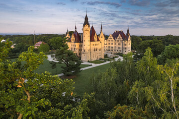 Majestic Moszna Castle on a warm summer morning, surrounded by lush greenery and glowing in soft golden sunlight, reflecting a fairytale-like ambiance perfect for travel and architecture themes.