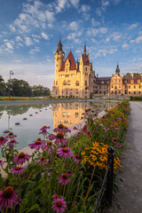 Majestic Moszna Castle on a warm summer morning, surrounded by lush greenery and glowing in soft golden sunlight, reflecting a fairytale-like ambiance perfect for travel and architecture themes.