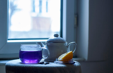 Butterfly pea flower tea turning purple from blue when lemon is added in a large sized glass cup on a small table with beige linen tablecloth in front of a window with copy space