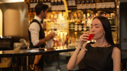 Beautiful guest looking at camera drinking at luxurious counter bar in mixed vodka Cosmopolitan red cocktail decorated with flower on bartender background in perfect atmosphere at nightclub. Vinosity.