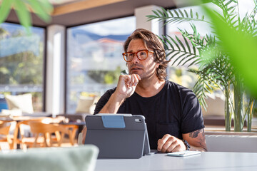 A man is sitting at a table in an office with a tablet working with a pensive expression