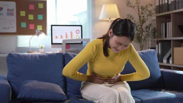 Female office worker experiencing intense abdominal pain while sitting on living room sofa, displaying signs of physical discomfort and health related distress