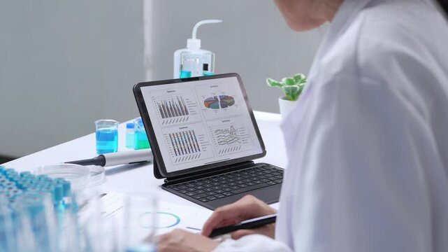 Professional female scientist in white coat analyzing data and charts on a tablet in a modern laboratory, surrounded by scientific equipment and blue liquids