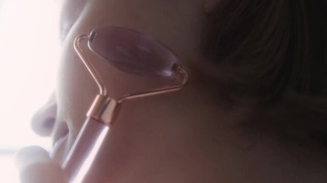 Close-up view of brunette girl using face roller gently massaging cheekbone during morning beauty routine