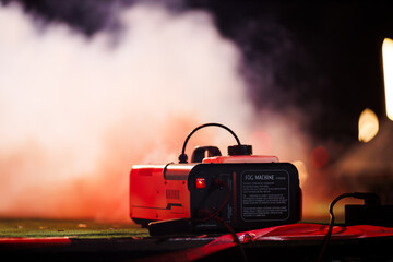 Close-up of a fog machine emitting thick smoke in a dramatic nighttime setting. The machine's glowing power switch and cables add a cinematic, atmospheric effect.