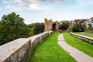 Two tourists are walking on a path towards Porta di Lavello, a medieval gate in Tuscania, Italy,...