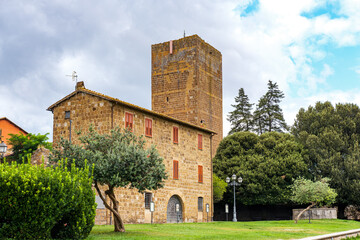 Torre di Lavello, a prominent medieval tower, rises above a historic building in Tuscania, Italy, surrounded by lush vegetation and showcasing the town's architectural heritage under a cloudy sky